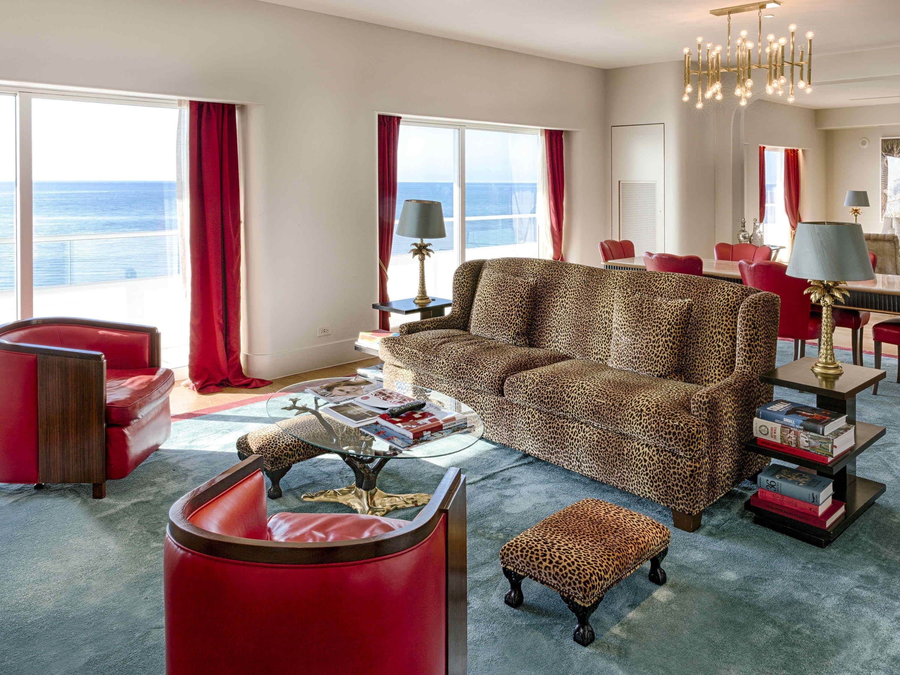 A modern living room with a leopard print sofa, red armchairs, a glass coffee table, and a view of the ocean.