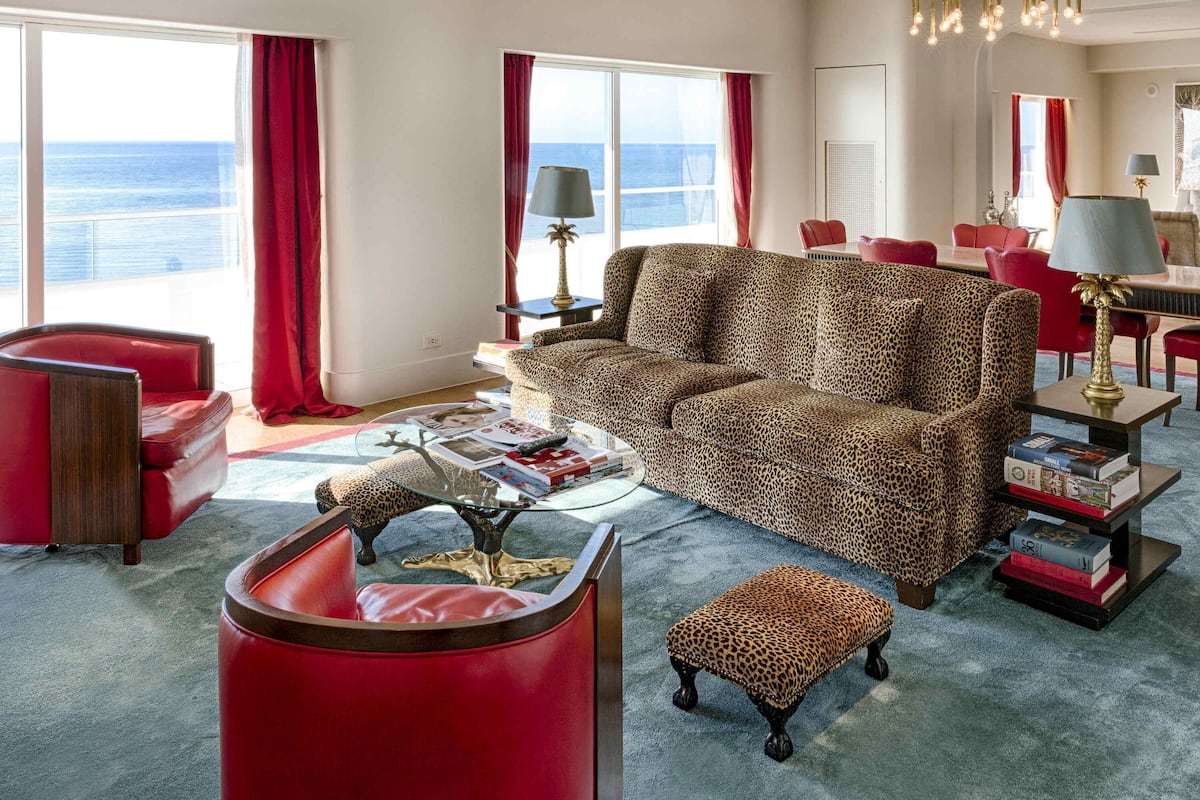 Ein modernes Wohnzimmer mit einem Couchgarnitur in Leopardenmuster, roten Sessel und einem Glastisch, mit Blick auf den Ozean.