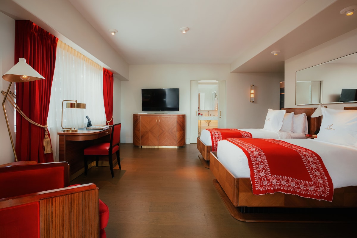 A hotel room with a wooden desk, a television, a bed with white bedding and a red throw, and a large mirror.