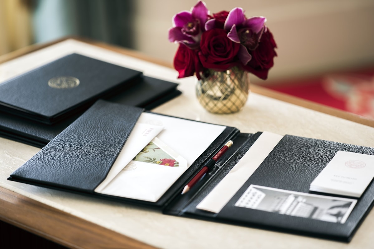 A desk with a black leather portfolio, a vase with purple flowers, and a pen.