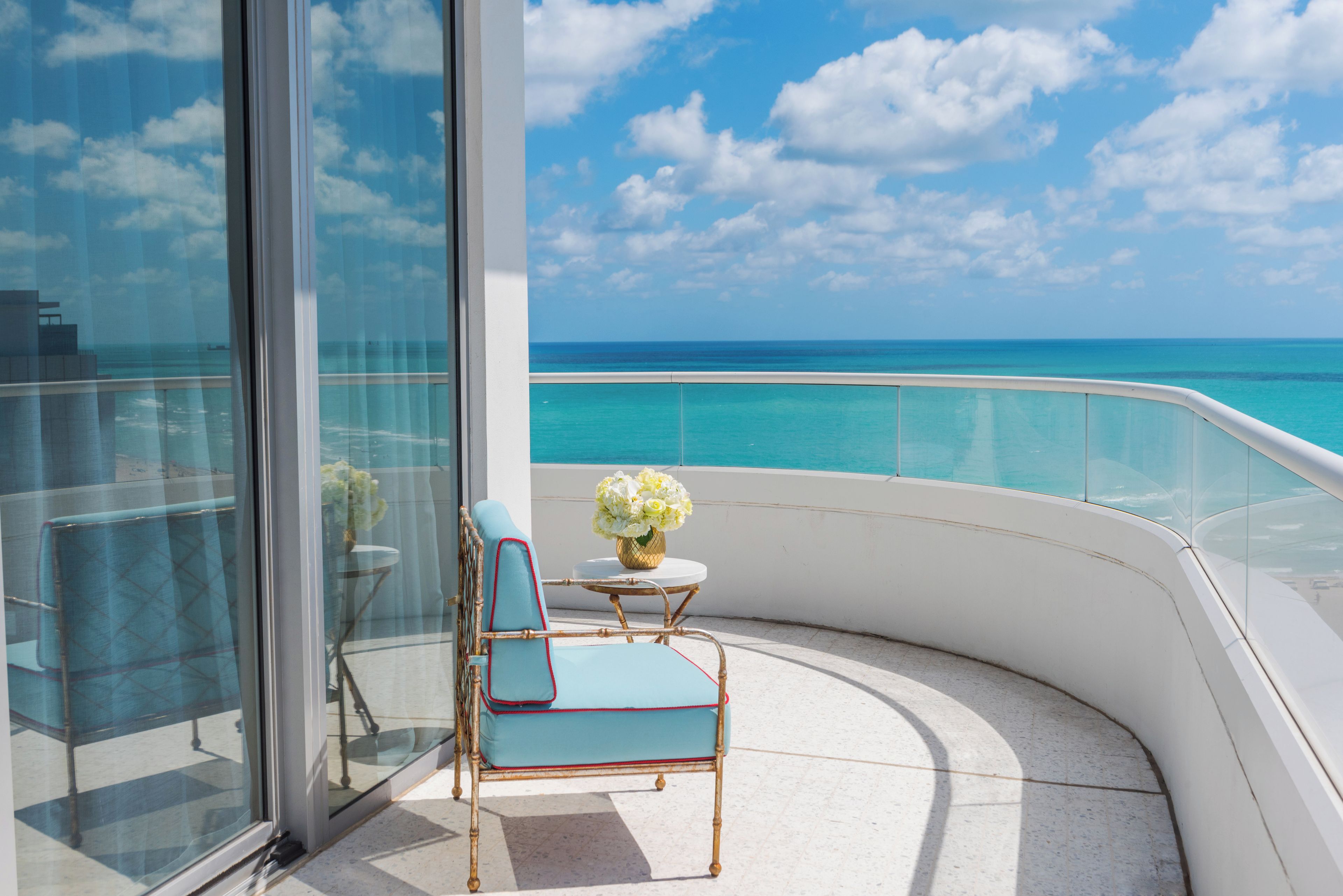 A balcony with a view of the ocean, a chair, a small table with a vase of flowers, and a glass railing.