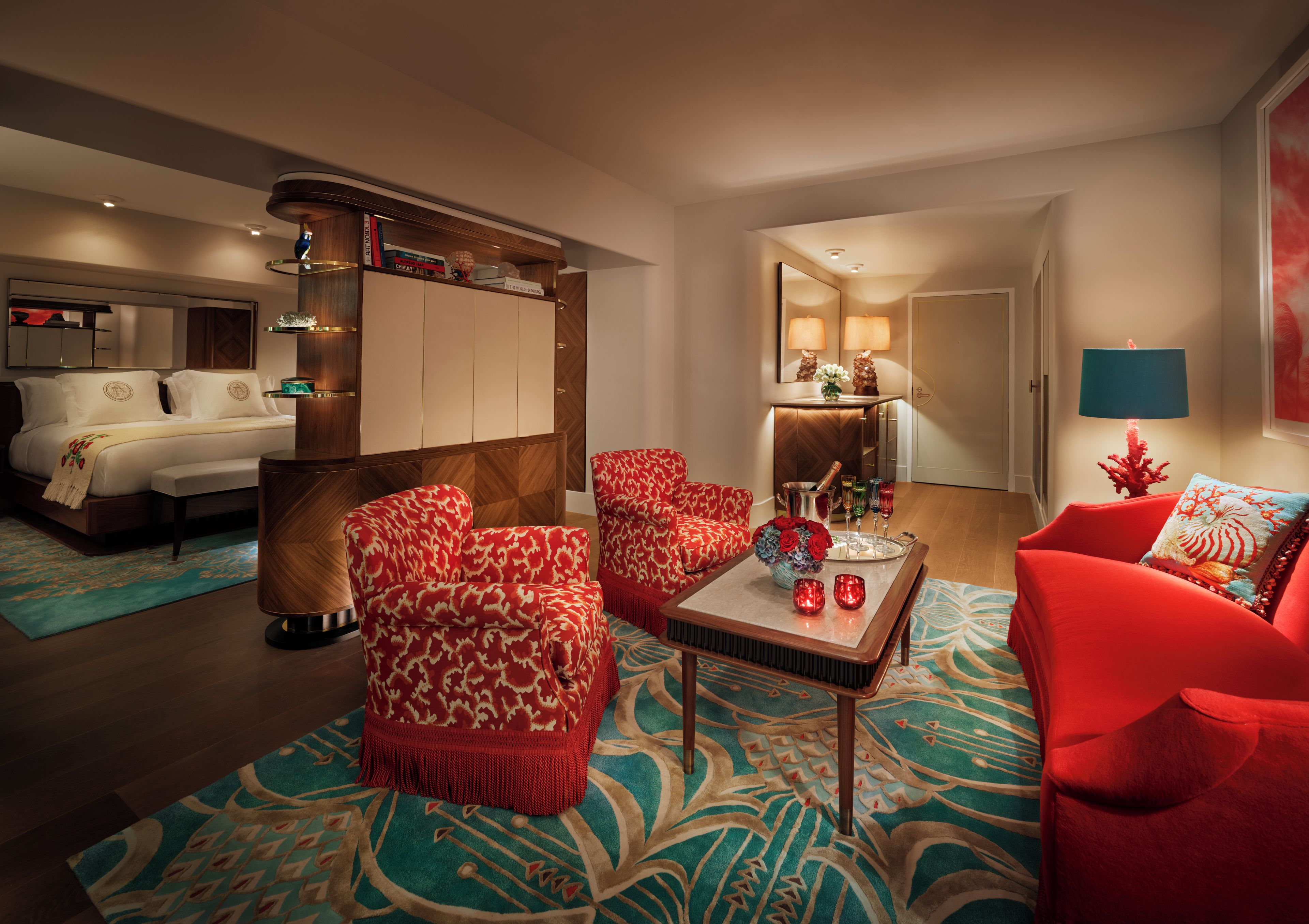 A hotel room with a bed, two red patterned armchairs, a coffee table with decorative items, a wooden cabinet, and a mirror.