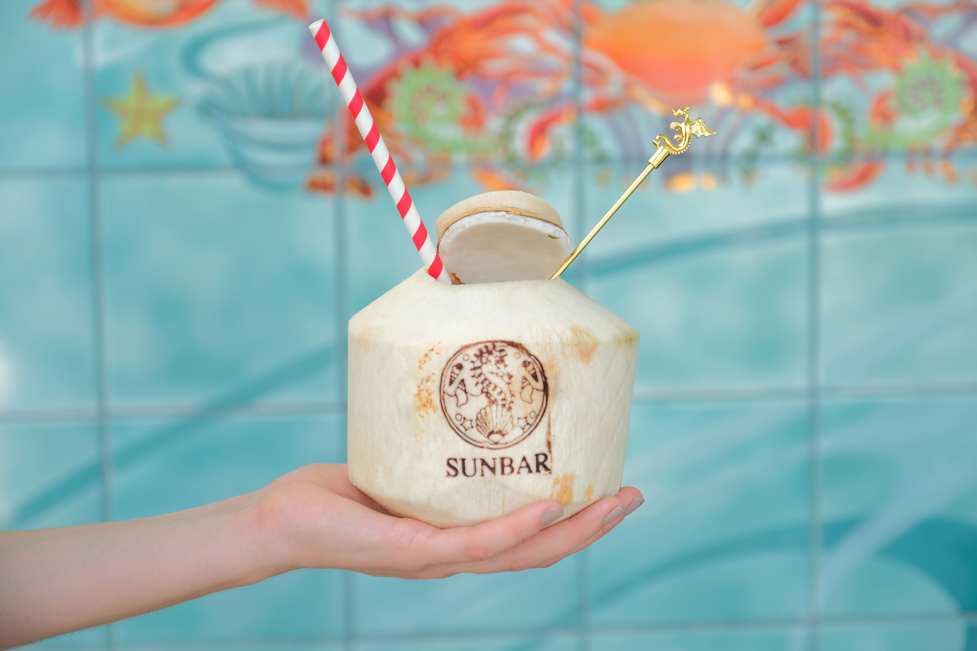 Una mano sosteniendo un coco con una pajita y una varita dorada, con la etiqueta 'SUNBAR'.