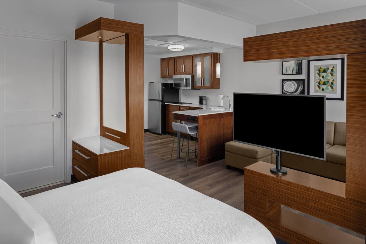 A modern hotel room with a bed, a TV, a kitchenette, and a dining area.