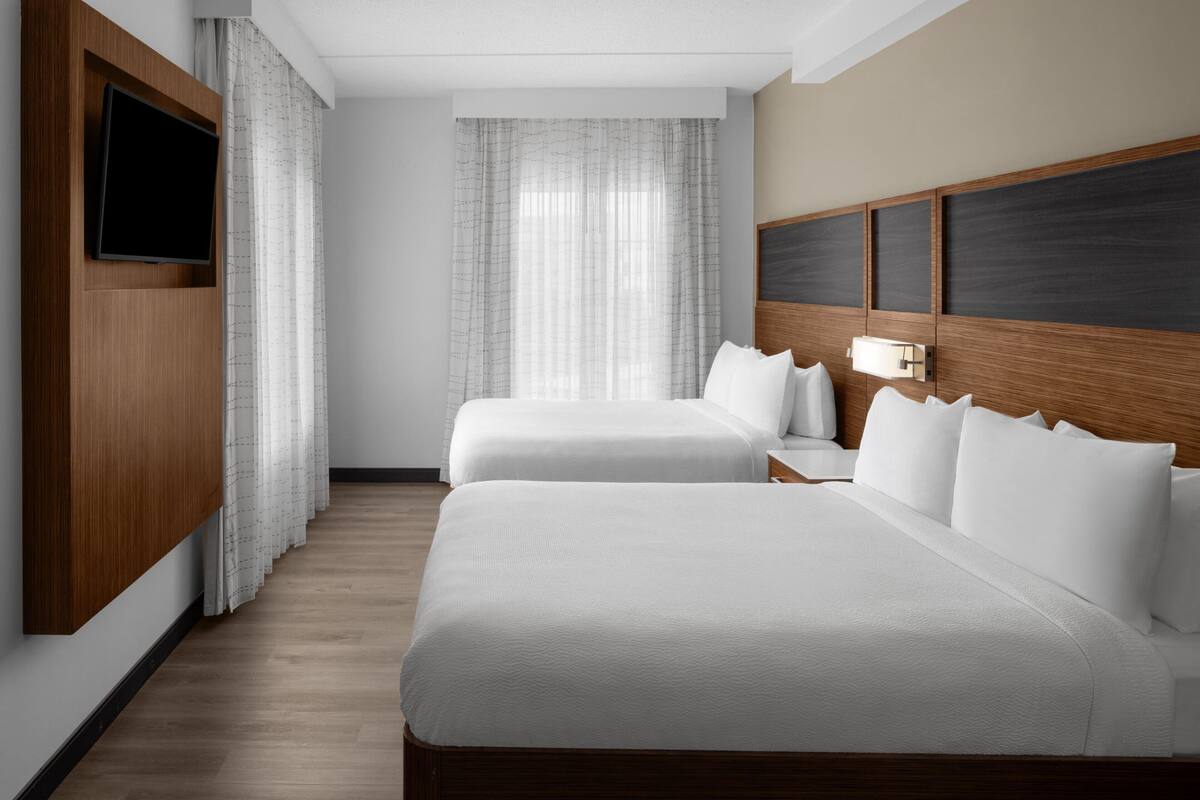 A hotel room with two beds, a flat-screen TV mounted on the wall, a wooden headboard, and a window with sheer curtains.