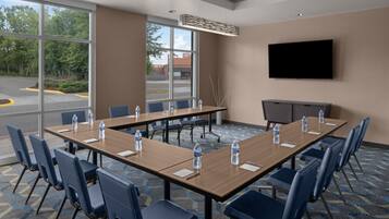 A conference room with a long table, blue chairs, water bottles, a flat-screen TV, and a large window with a view of a parking lot and trees.