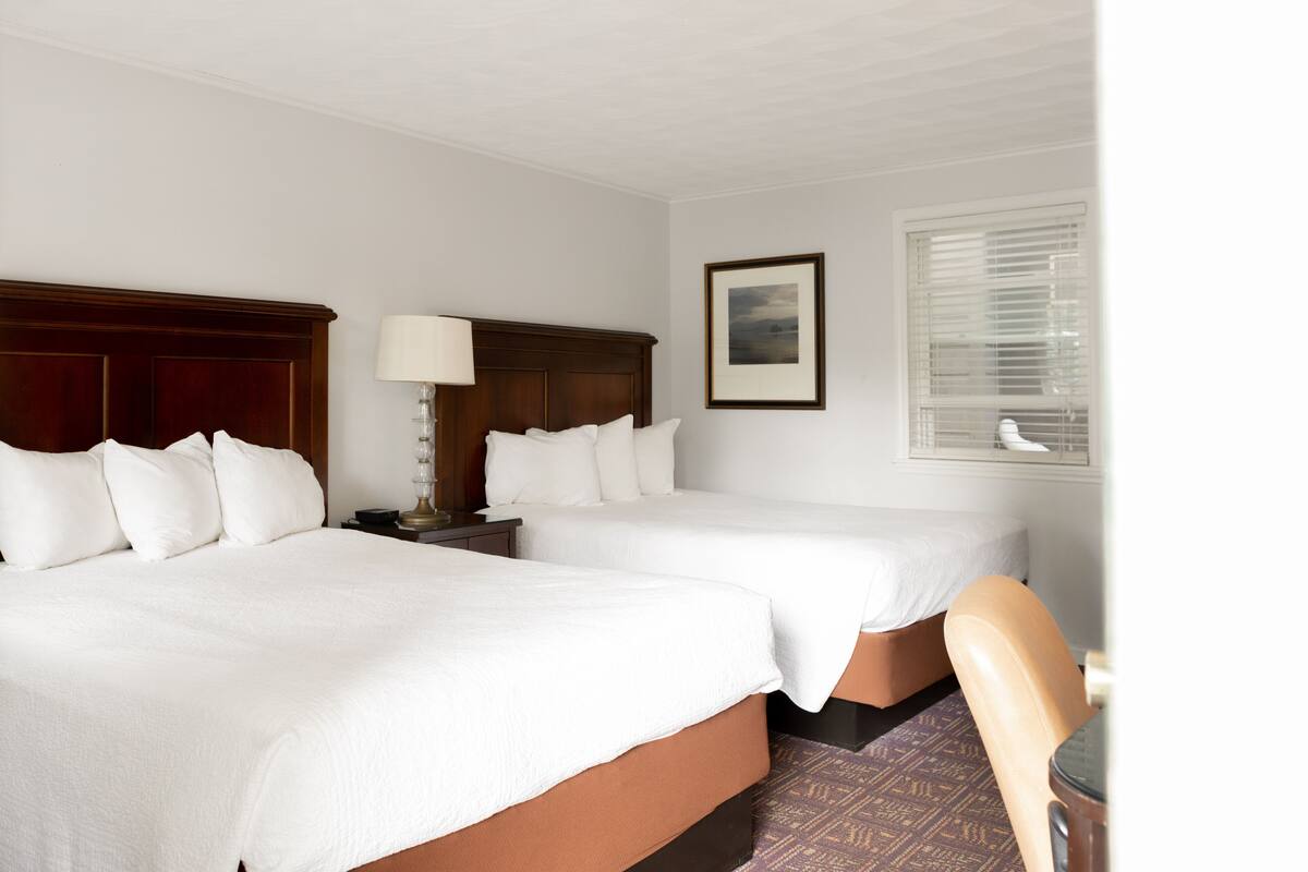 Habitación de hotel con dos camas, una mesita de noche con lámpara, un cuadro en la pared y una ventana con persianas.