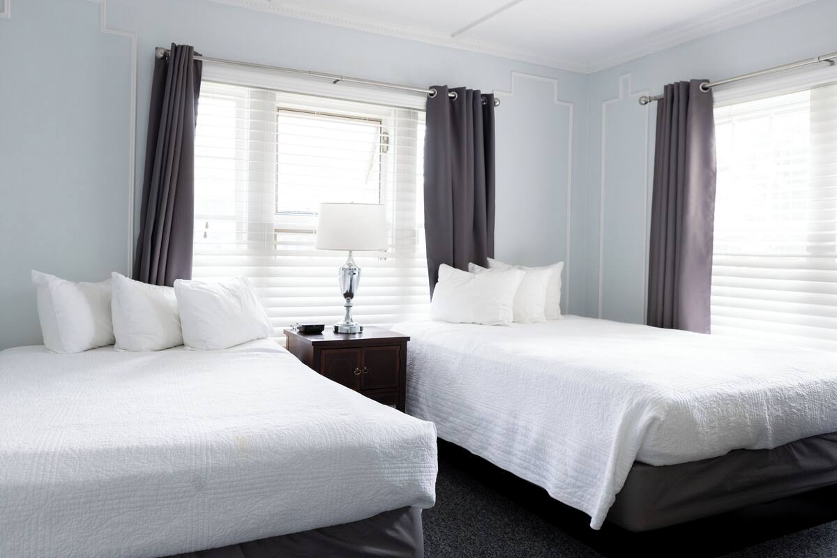 Habitación de hotel con dos camas, una mesita de noche, una lámpara y una ventana con cortinas.