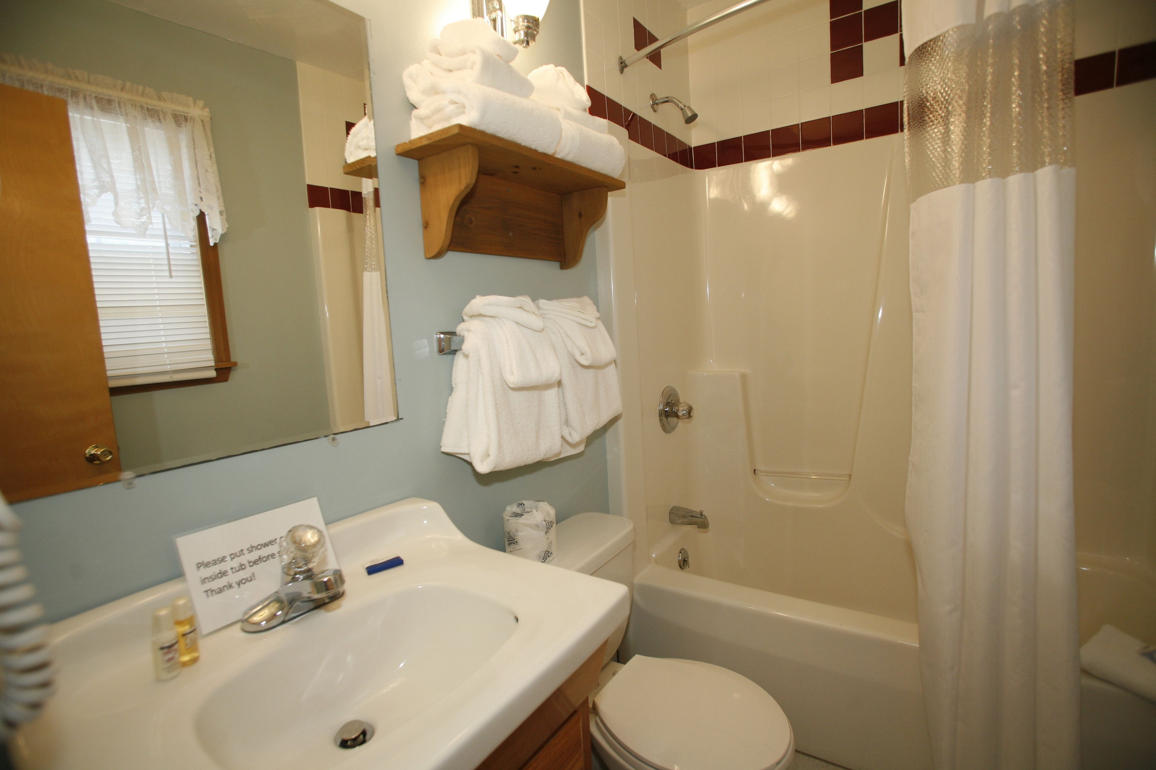 A bathroom with a sink, toilet, and bathtub. There are towels on a shelf and a sign instructing to put the shower curtain inside the tub before use.
