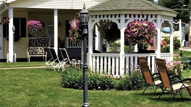 A white building with a covered porch and a gazebo with flower baskets. - Heritage of Lake George (Lake George)