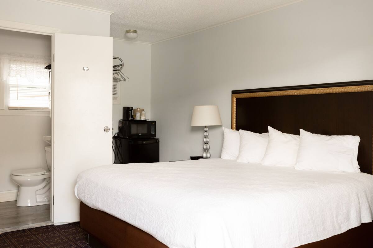 Habitación de hotel con una cama grande, una mesita de noche con lámpara, un escritorio pequeño, un espejo y un baño con inodoro y lavamanos.