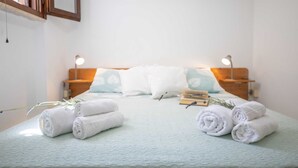 Una cama bien hecha con toallas blancas enrolladas y una ramita de romero, una mesita de noche de madera con una lámpara, y un cuadro colgado en la pared.