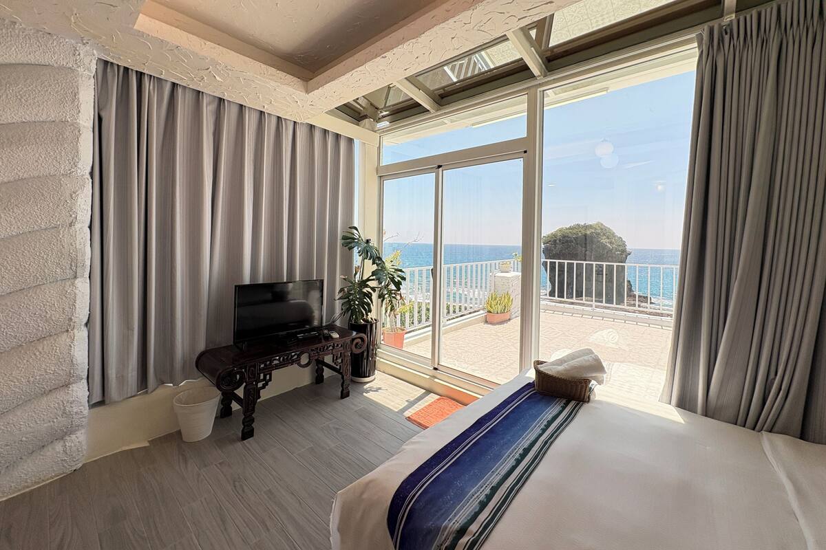 A bedroom with a large bed, a TV, a plant, and a balcony with a view of the sea.