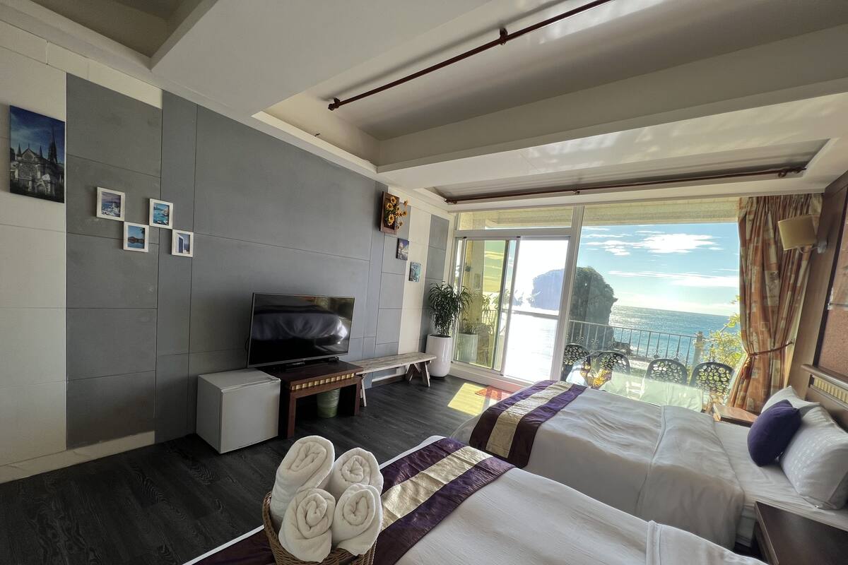 A hotel room with two beds, a TV, a small desk, and a balcony with a view of the sea.