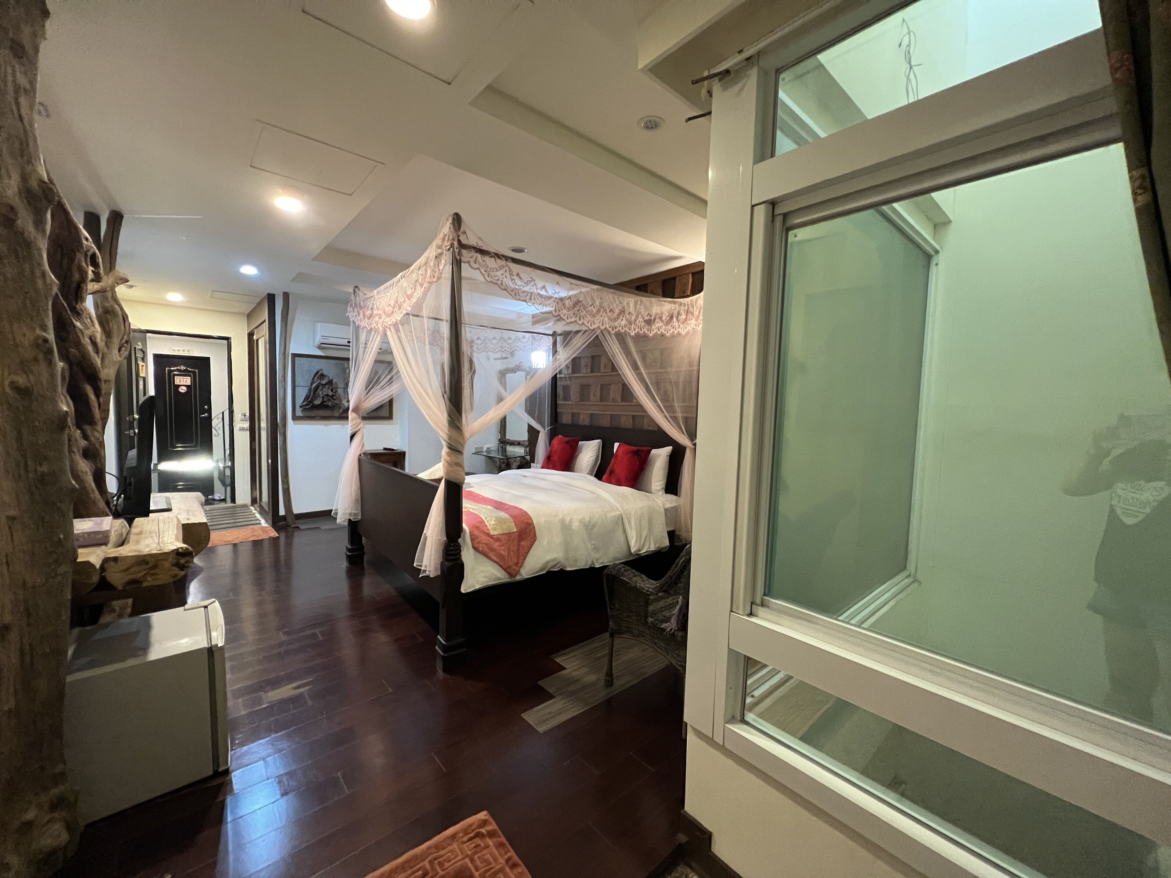 A bedroom with a four-poster bed, a large window, and a wooden floor.