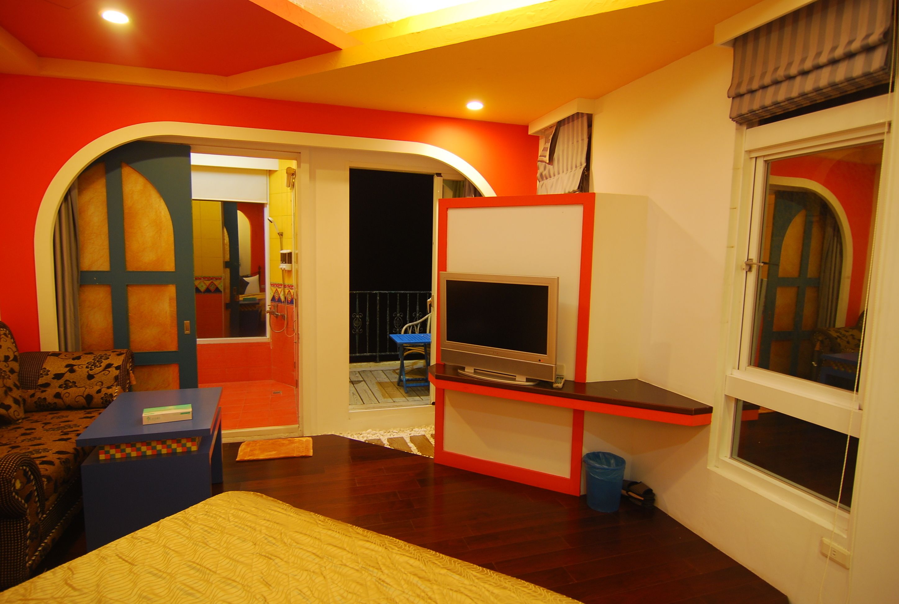 A room with a bed, a TV on a stand, a small table, and a chair. There is a balcony with a railing and a chair outside.