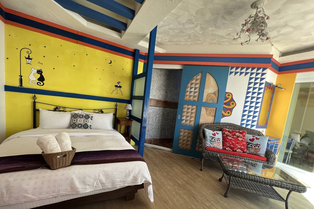 A room with a bed, a small table, and a wicker chair.