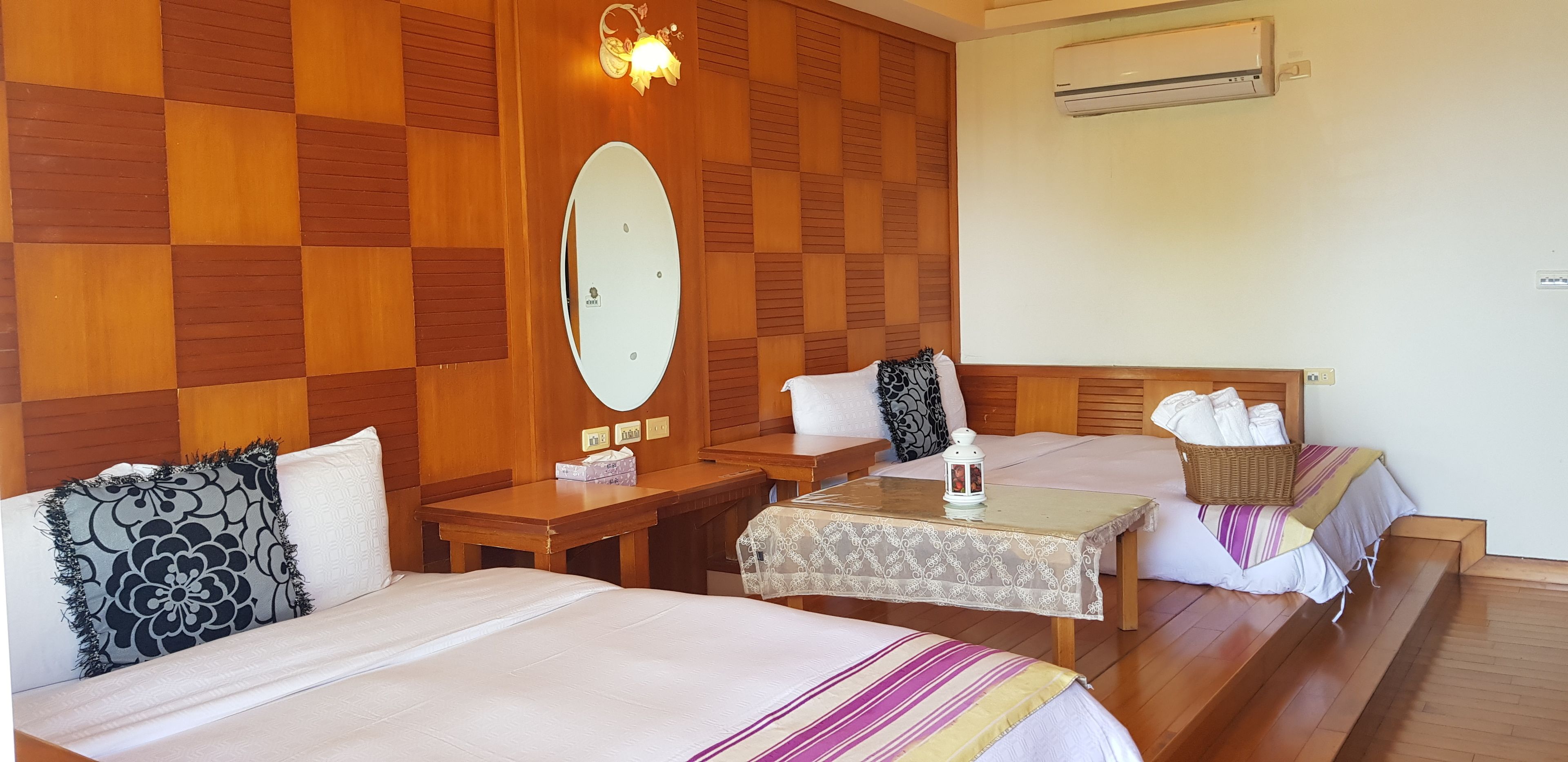 A hotel room with two beds, a wooden wall panel, a mirror, a table with a lantern, and an air conditioning unit.