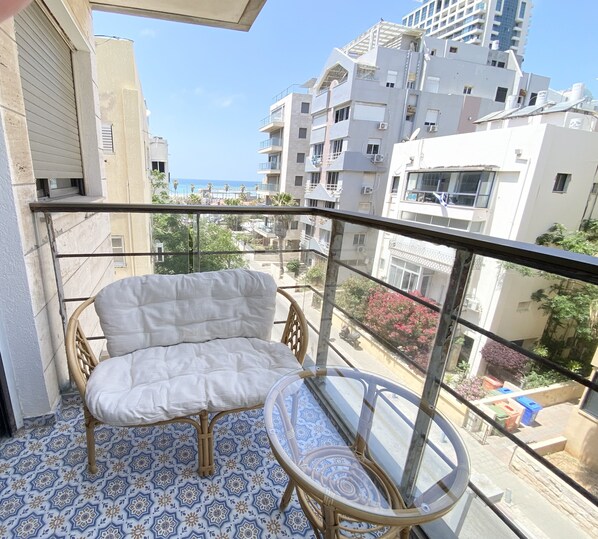 A balcony with a wicker chair and a small table, overlooking a residential area with buildings and a clear sky. - Sea Suites Apartments (Tel Aviv)