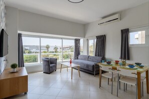 A modern living room with a grey sofa, a wooden coffee table, and a dining table with chairs. There is a TV on a stand and a small plant on the coffee table. - Sea Suites Apartments (Tel Aviv)