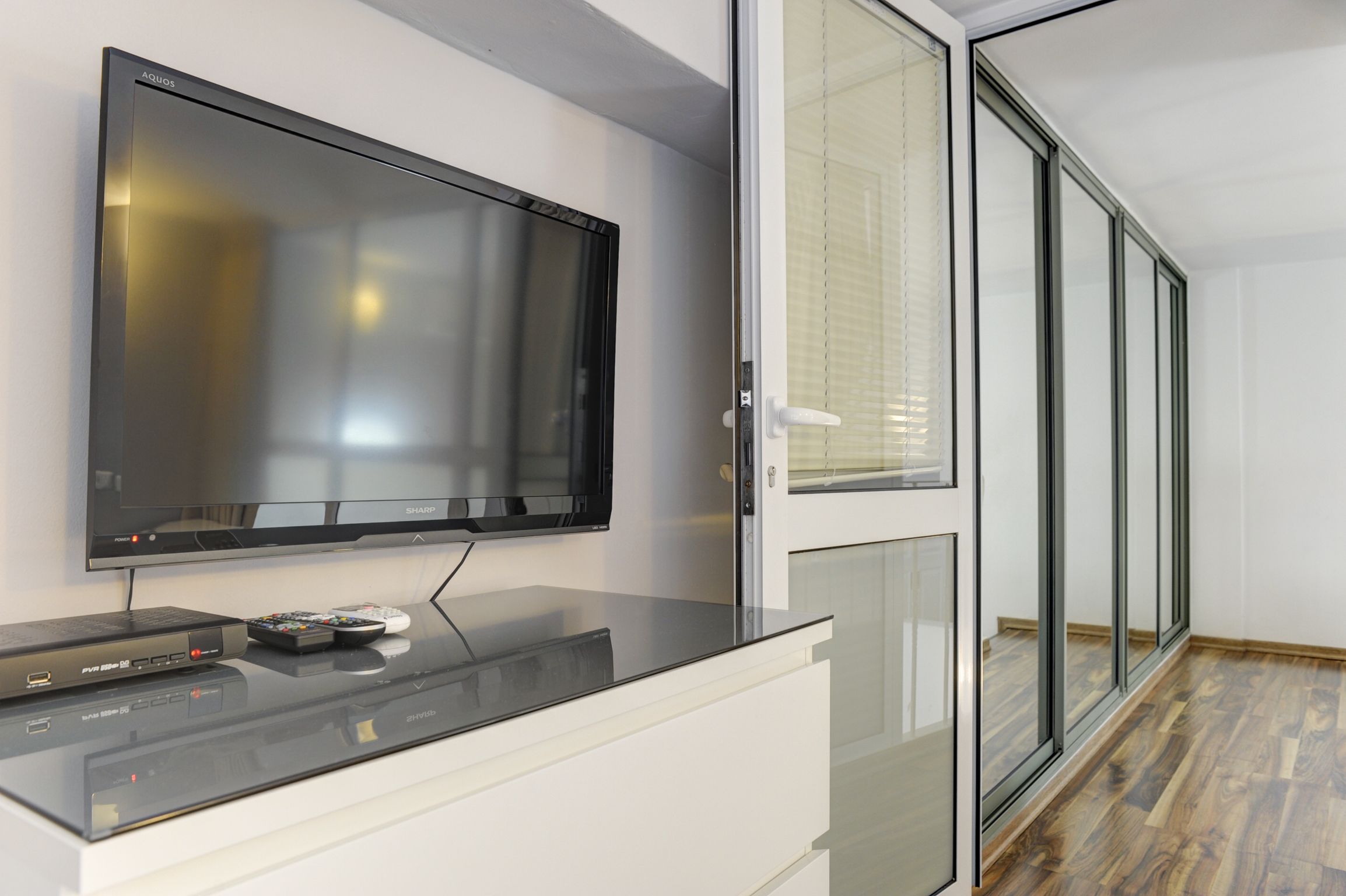 Un salon moderne avec un téléviseur à écran plat fixé au mur, une table basse en verre et une porte-fenêtre coulissante donnant sur un espace doté d’un plancher en bois.