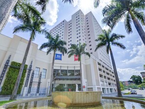 A modern building with a prominent 'ibis budget' sign, a central courtyard with a fountain, and palm trees surrounding the area. - ibis Rio de Janeiro Shopping Nova America (Rio de Janeiro)