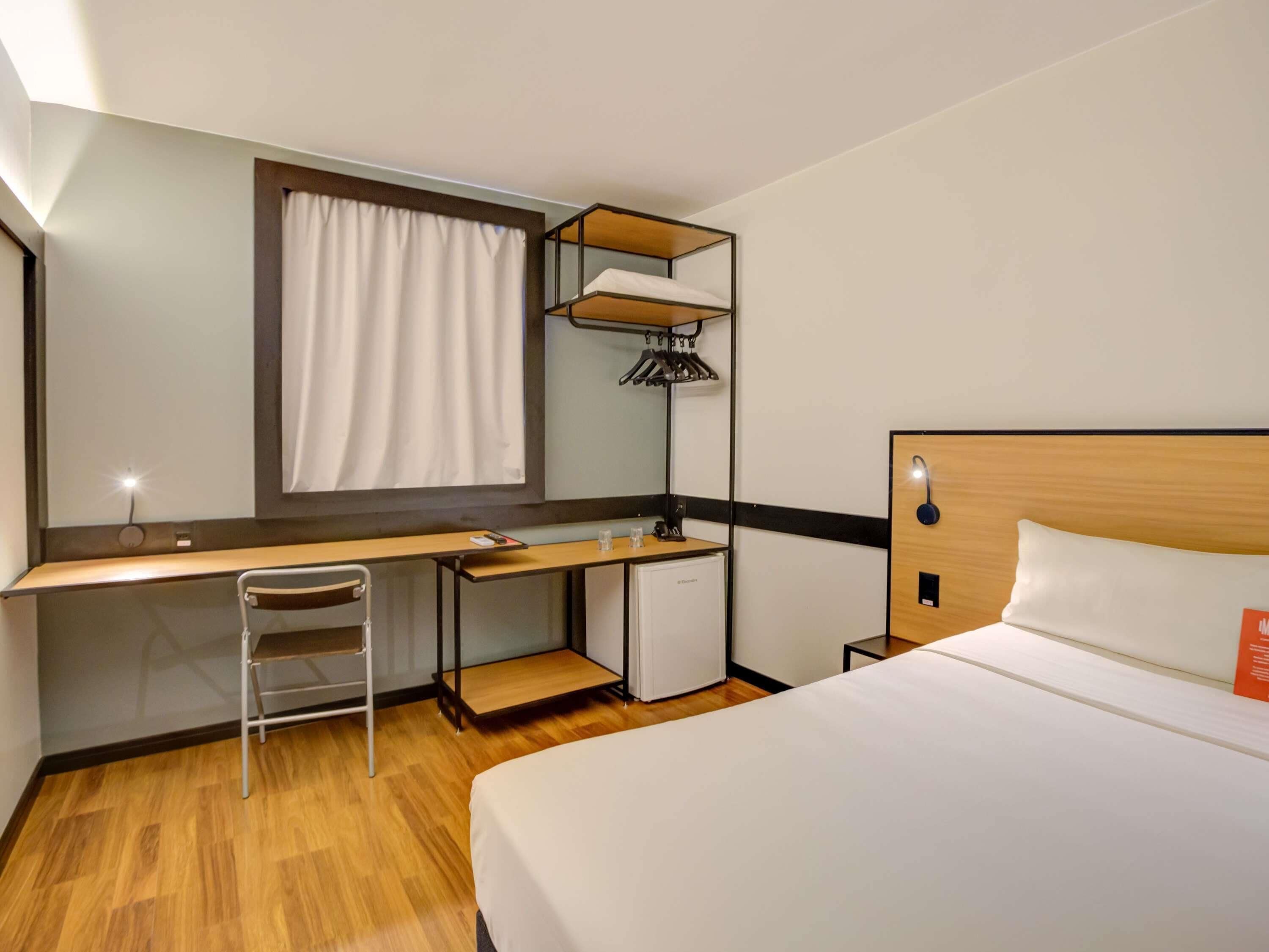 A hotel room with a bed, desk, chair, and a small kitchenette.