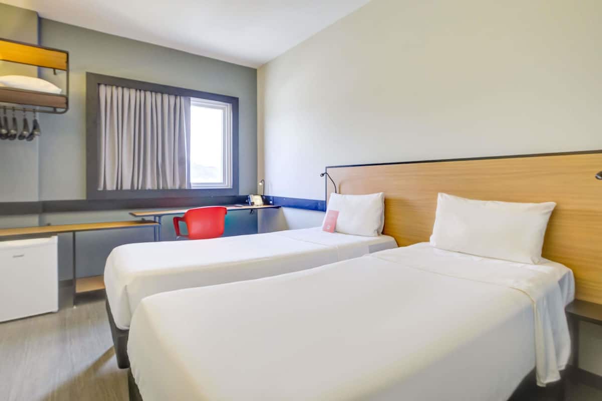 Ein Hotelzimmer mit zwei Betten, einem Schreibtisch, einem Stuhl, einem Fenster mit Vorhängen und einer Wandleuchte.