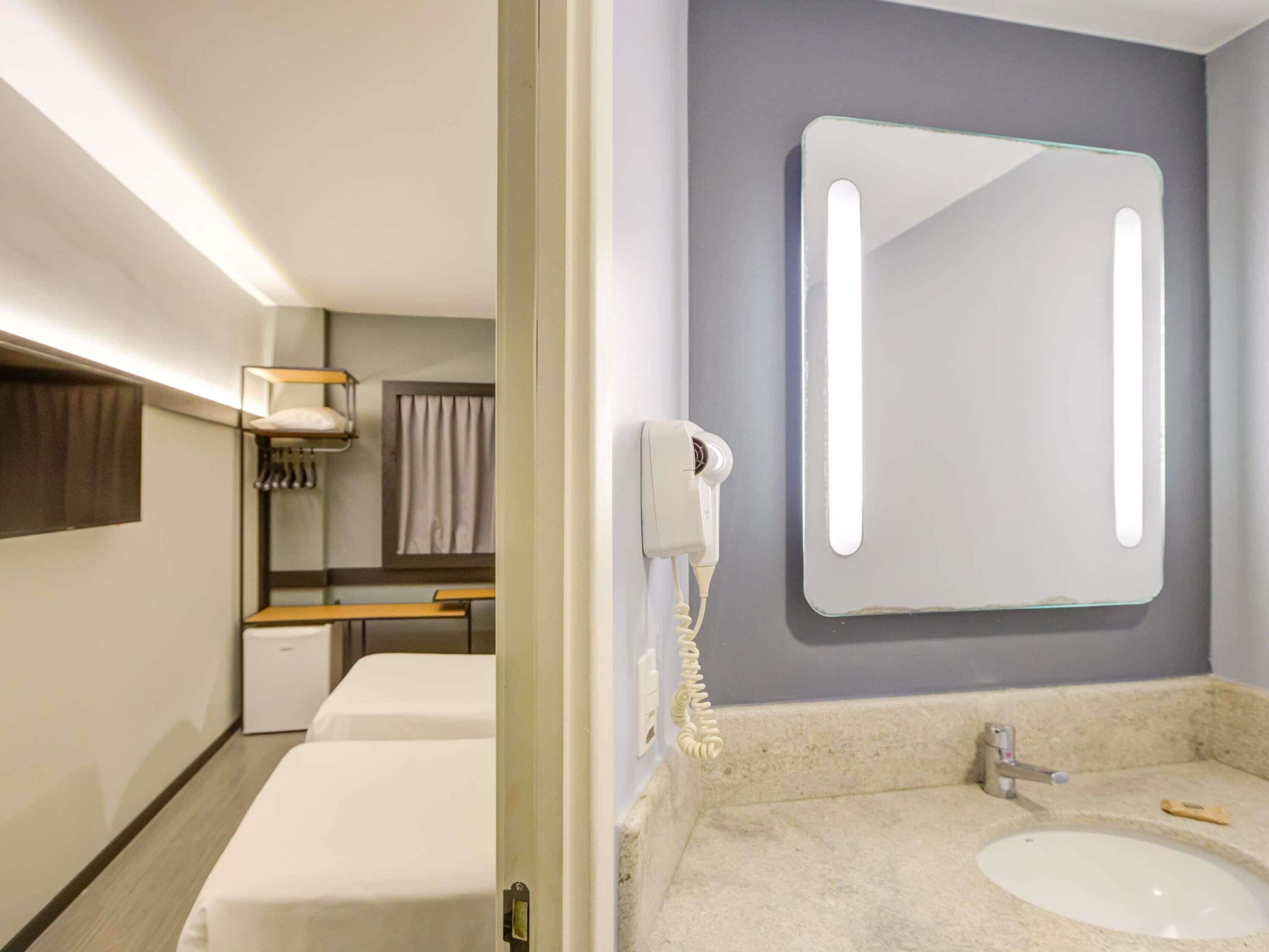 A hotel room with a bed, a bedside table, a mirror with a light, a phone, and a bathroom with a sink and mirror.