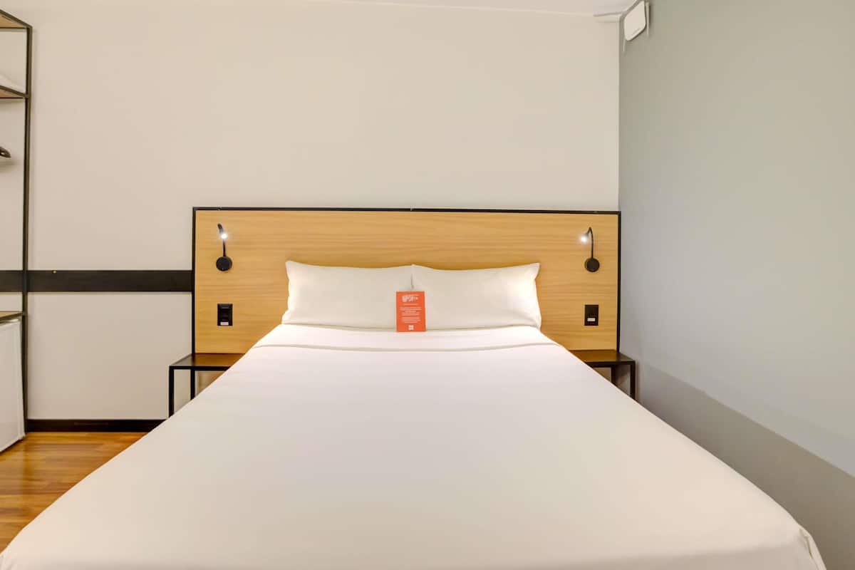 Ein ordentlich bezogenes Bett in einem Hotelzimmer mit einem hölzernen Kopfteil, zwei Nachttischen und einer wandmontierten Lampe.