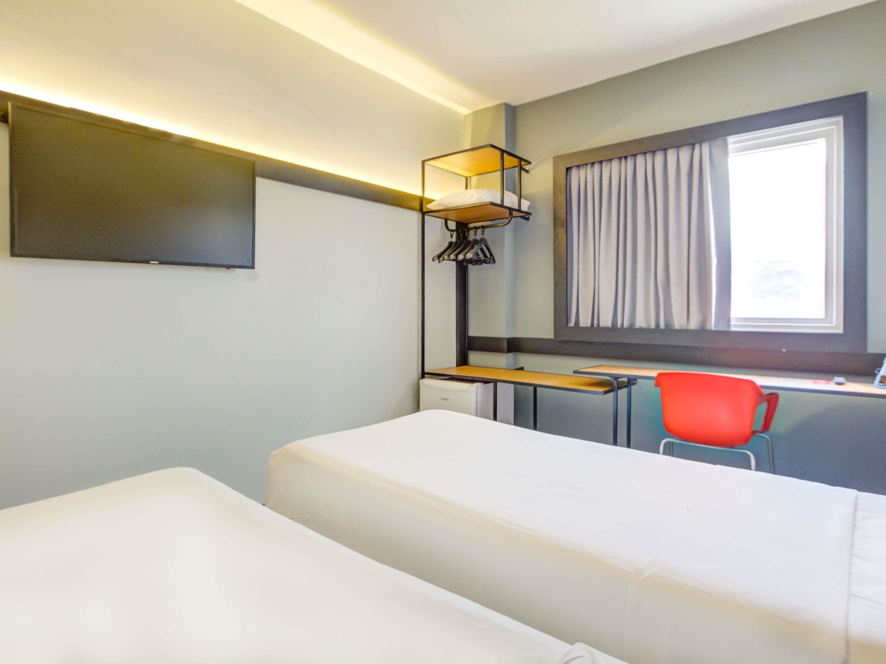 Habitación de hotel con cama, escritorio, silla, televisor y ventana con cortinas.