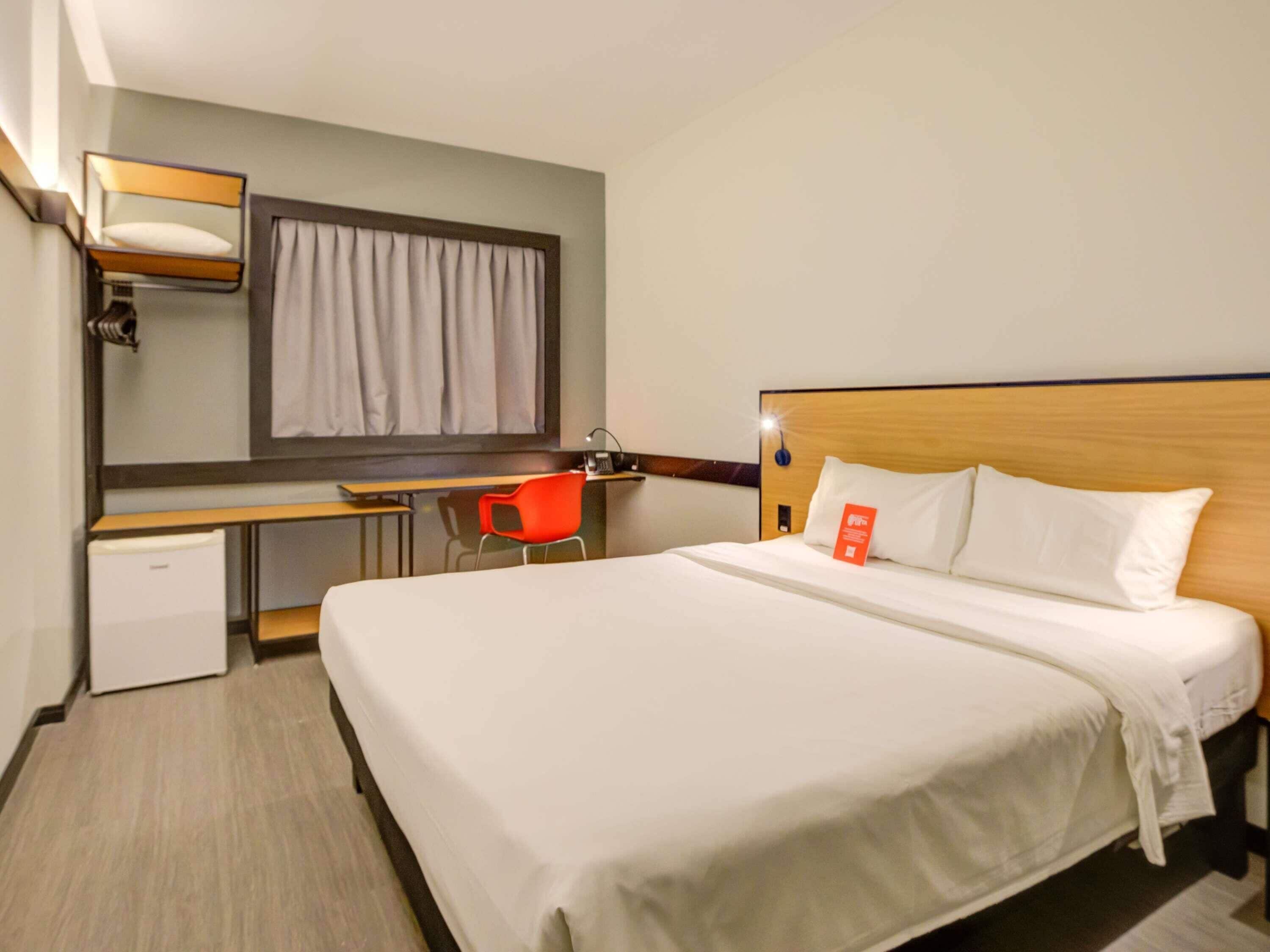 A hotel room with a single bed, a desk with a chair, a small fridge, and a window with curtains.