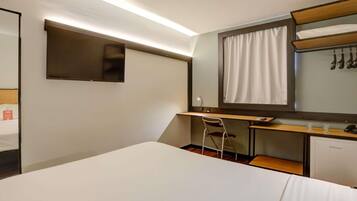 Una habitación de hotel con cama, escritorio, silla, televisor y espejo.