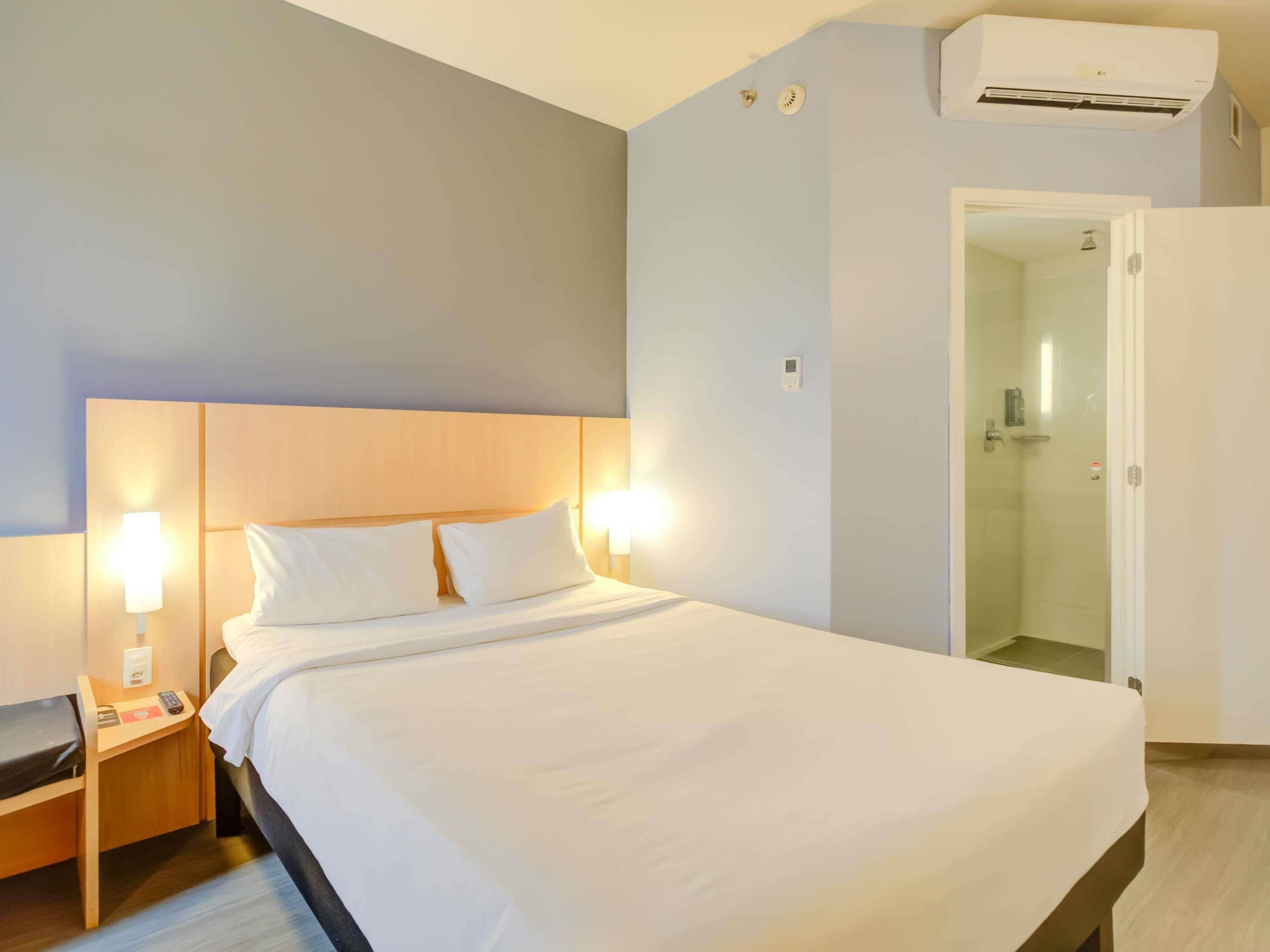 Una habitación de hotel con cama, lámparas de noche, un equipo de aire acondicionado y una puerta que da a un baño.