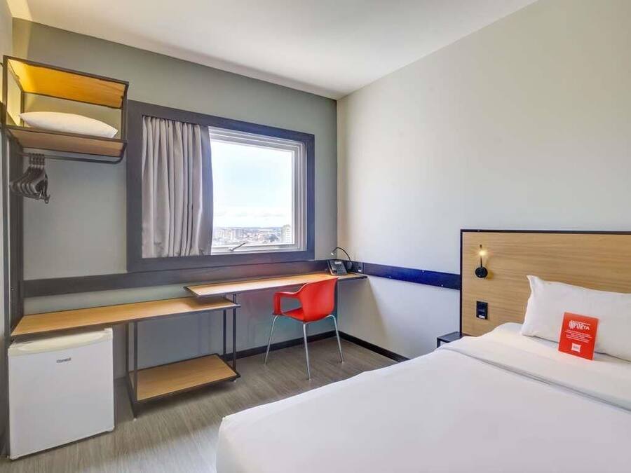 Ein Hotelzimmer mit Bett, Schreibtisch, Stuhl, kleinem Kühlschrank, Fenster mit Vorhängen und Nachttisch mit Lampe.