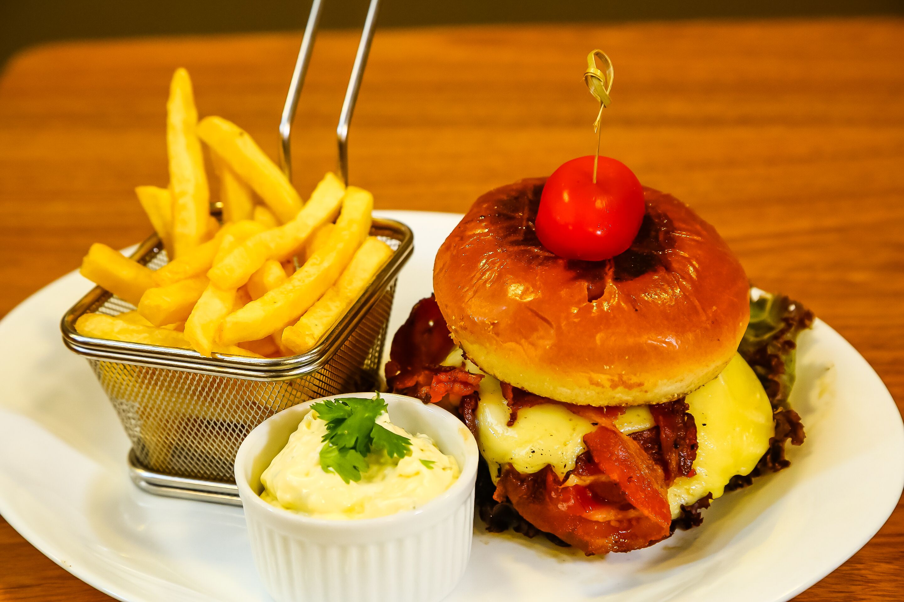 A burger with a cherry on top, French fries, and a small bowl of sauce on a white plate.