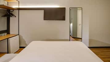 Habitación de hotel minimalista con cama, un pequeño escritorio, un espejo y televisión montada en la pared.