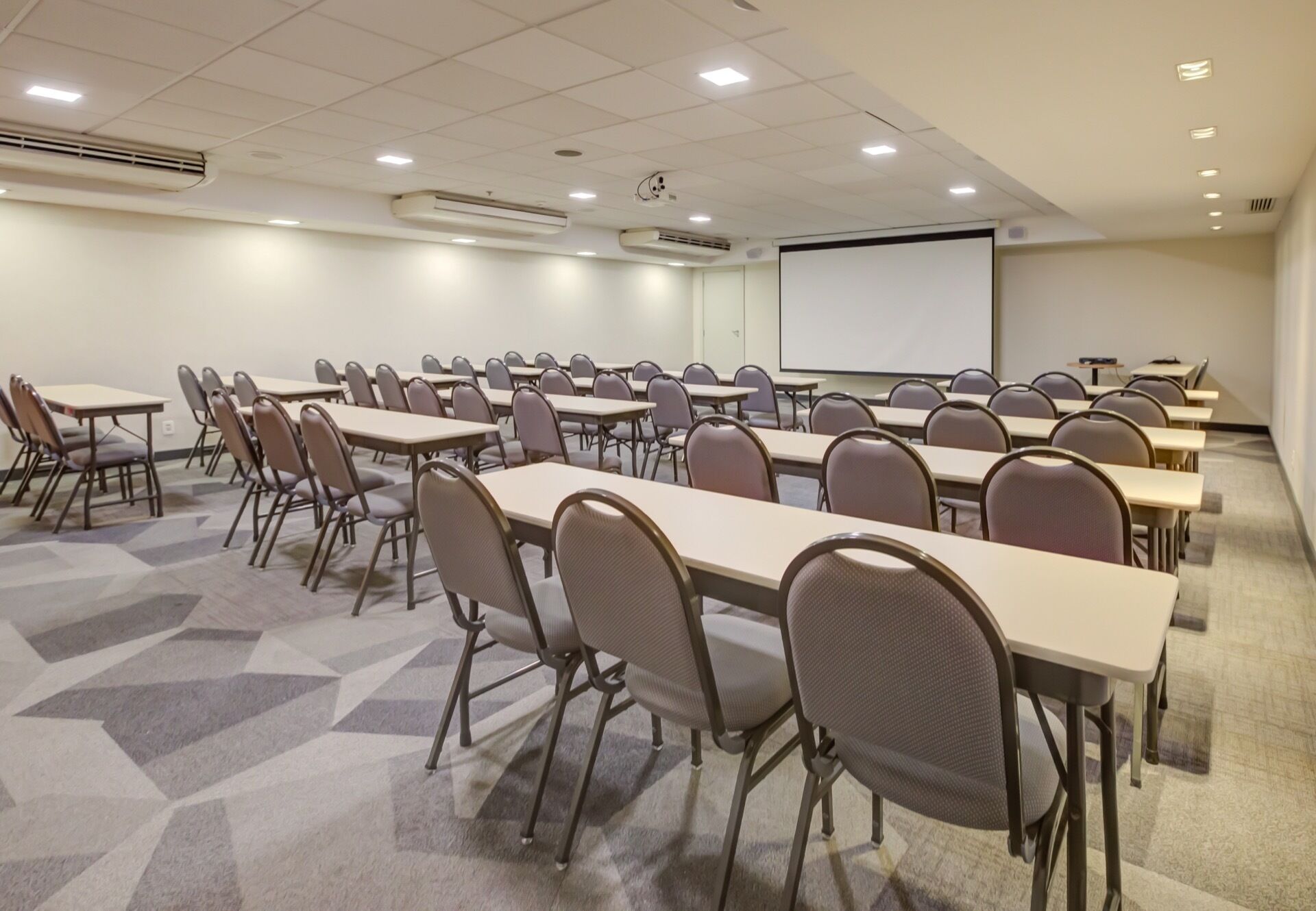Uma sala de conferências com mesas e cadeiras dispostas em fileiras, uma tela de projetor e aparelhos de ar condicionado.