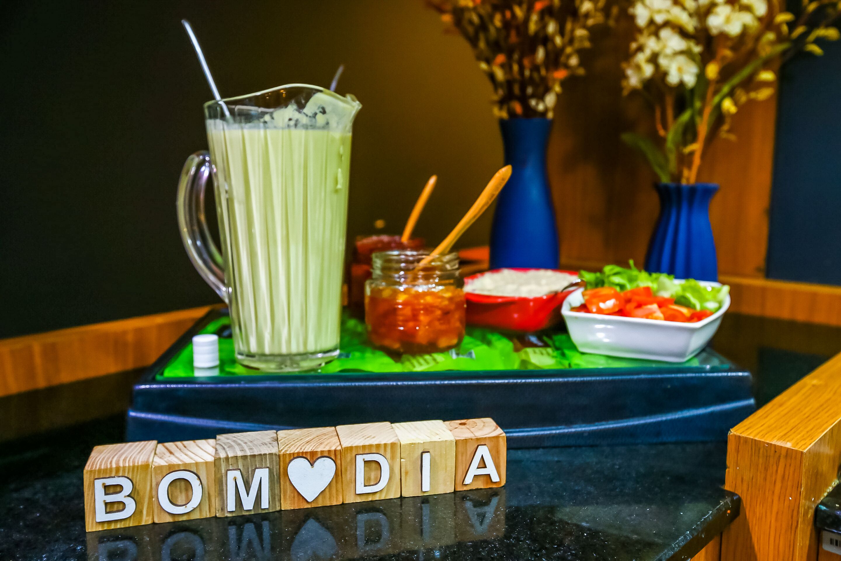 A table with a green placemat, a glass of green beverage, a bowl of salad, and wooden blocks spelling "BOM DIA".