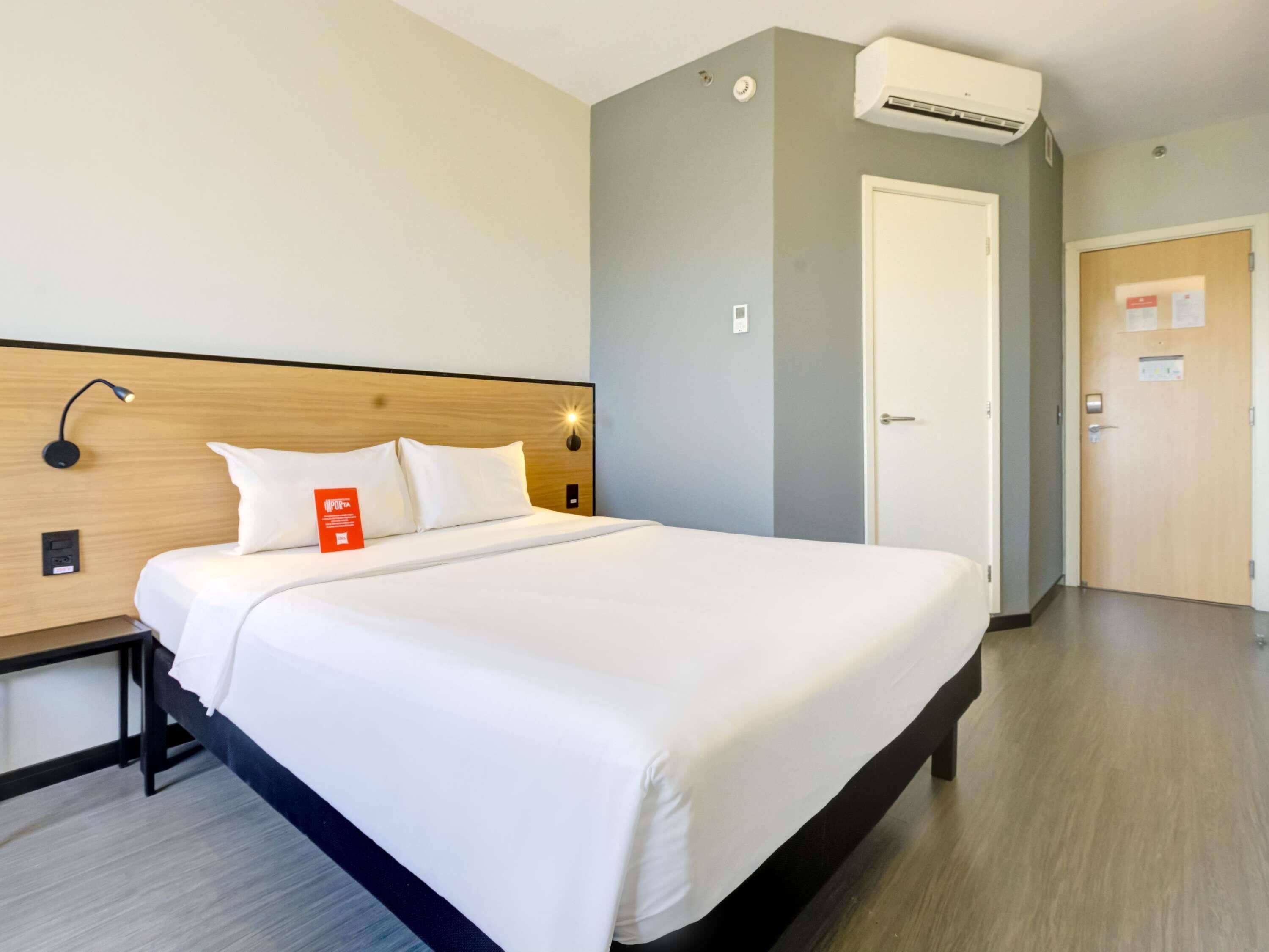 Quarto de hotel com cama, mesa de cabeceira, luminária, aparelho de ar condicionado e porta.