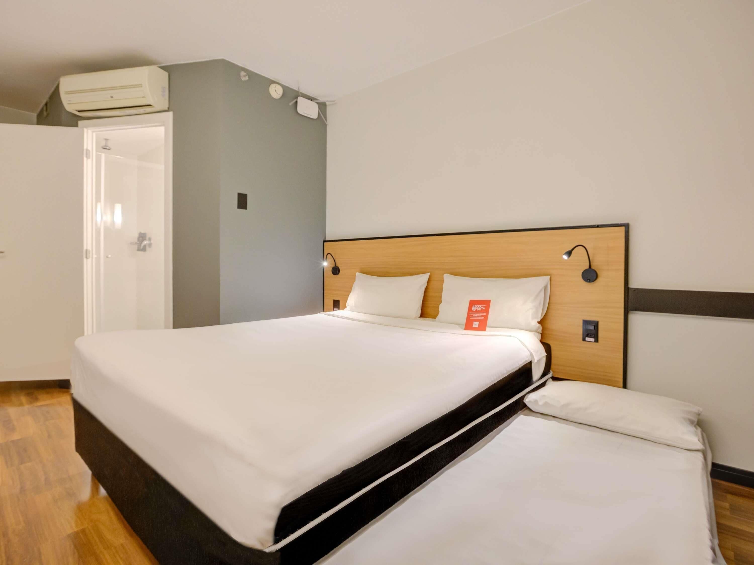 A hotel room with a bed, a wall-mounted air conditioner, a clock, and a door.