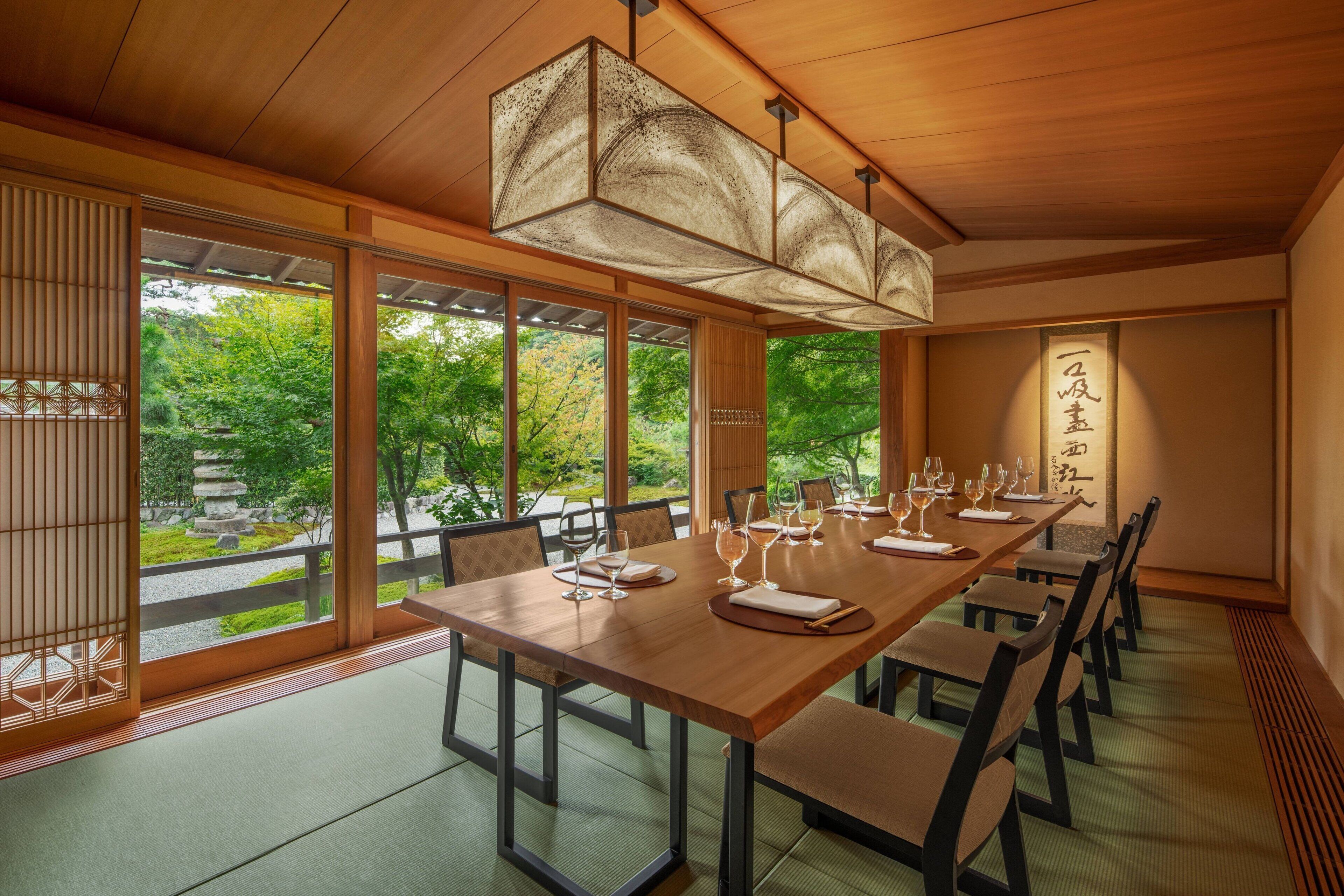 A traditional Japanese-style dining room with a long wooden table set for a meal, large windows offering a view of a garden, and a hanging light fixture.