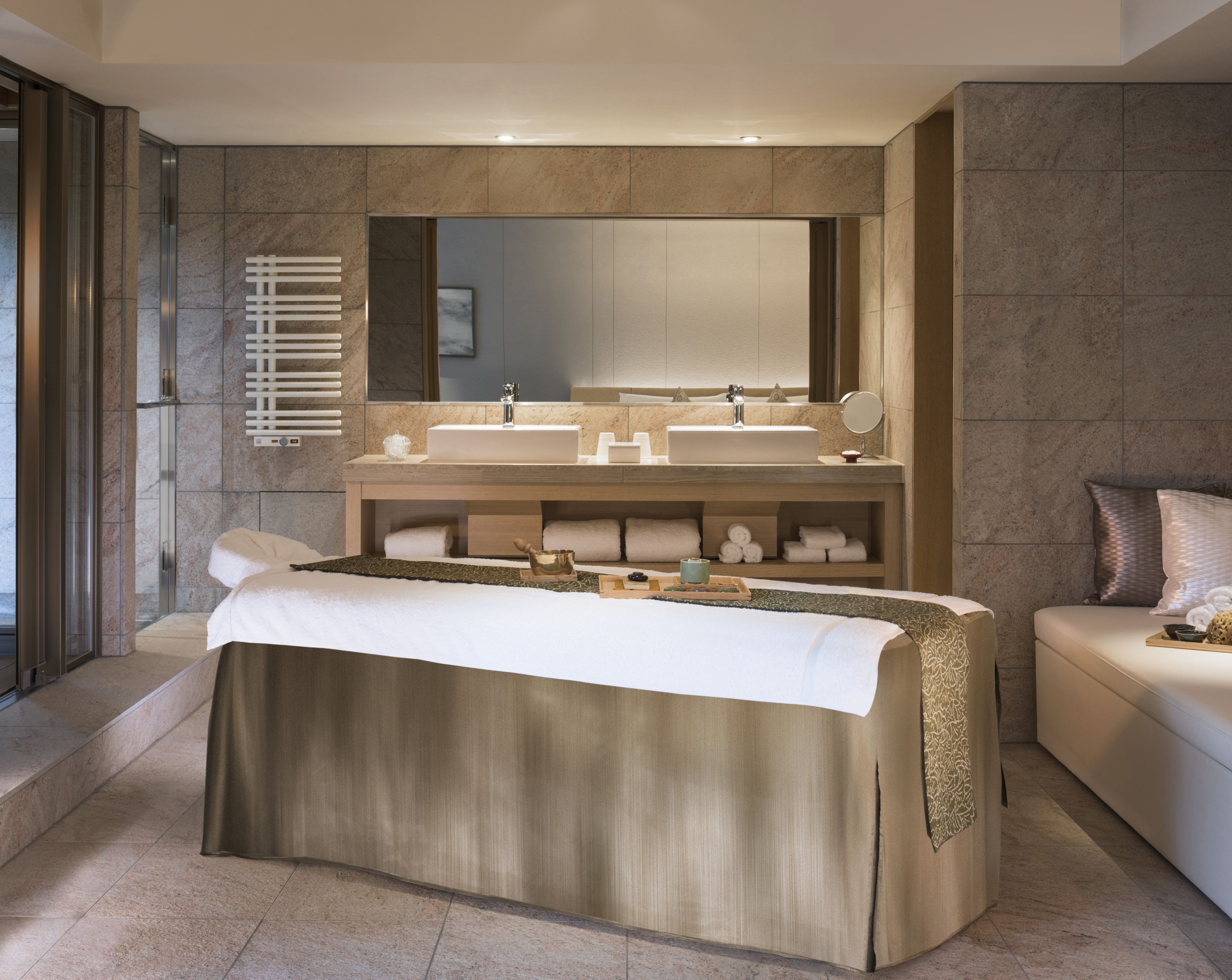 A modern bathroom with a large mirror, two sinks, a heated towel rail, and a bed with a large towel.
