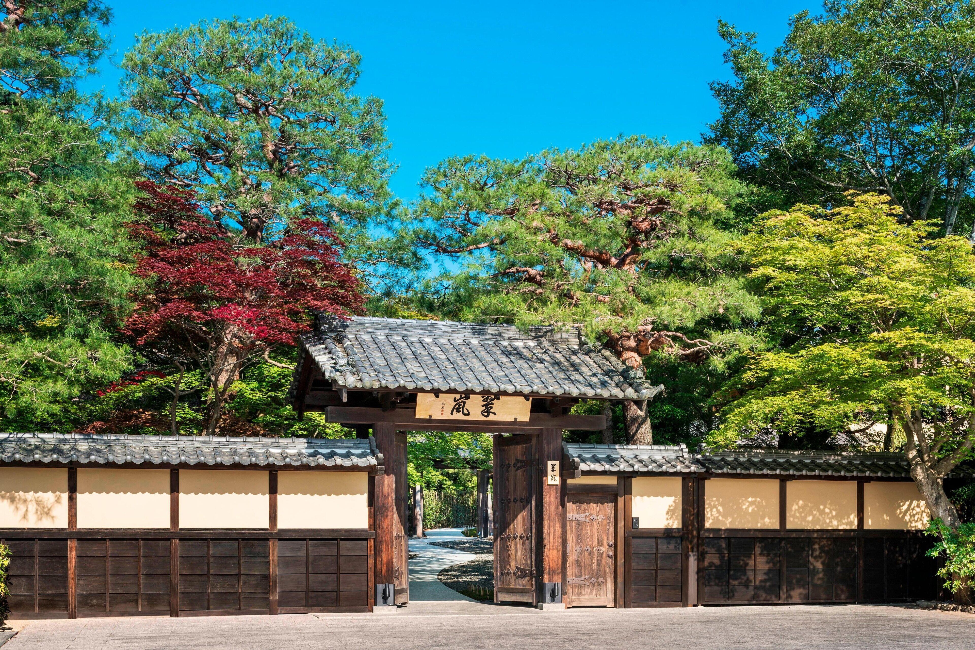 A traditional Japanese gate with a wooden structure and a tiled roof, surrounded by lush greenery and trees.