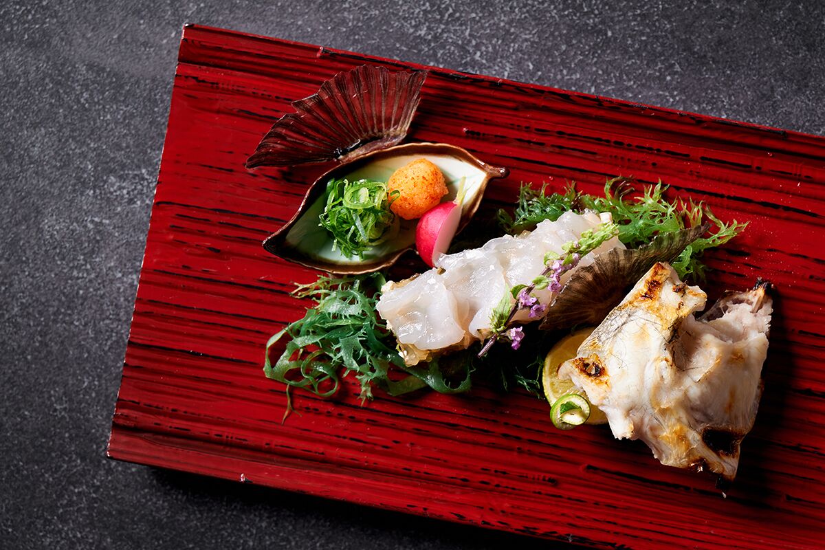 Una mesa de madera roja con un plato de comida que incluye pescado, verduras de hoja verde y una pequeña concha.
