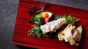 Una mesa de madera roja con un plato de comida que incluye pescado, verduras de hoja verde y una pequeña concha.