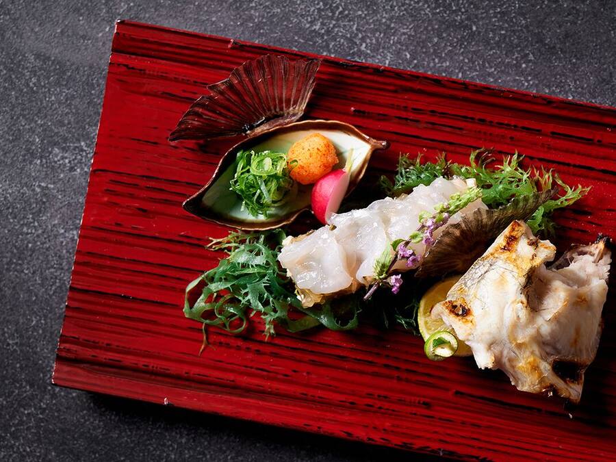 Una mesa de madera roja con un plato de comida que incluye pescado, verduras de hoja verde y una pequeña concha.