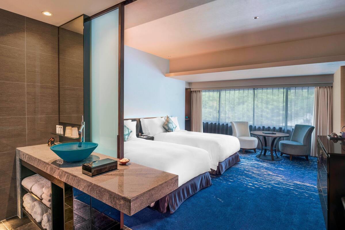 A hotel room with a large bed, a sitting area with a table and chairs, a large window with curtains, a bathroom with a large mirror and a sink with a blue basin.
