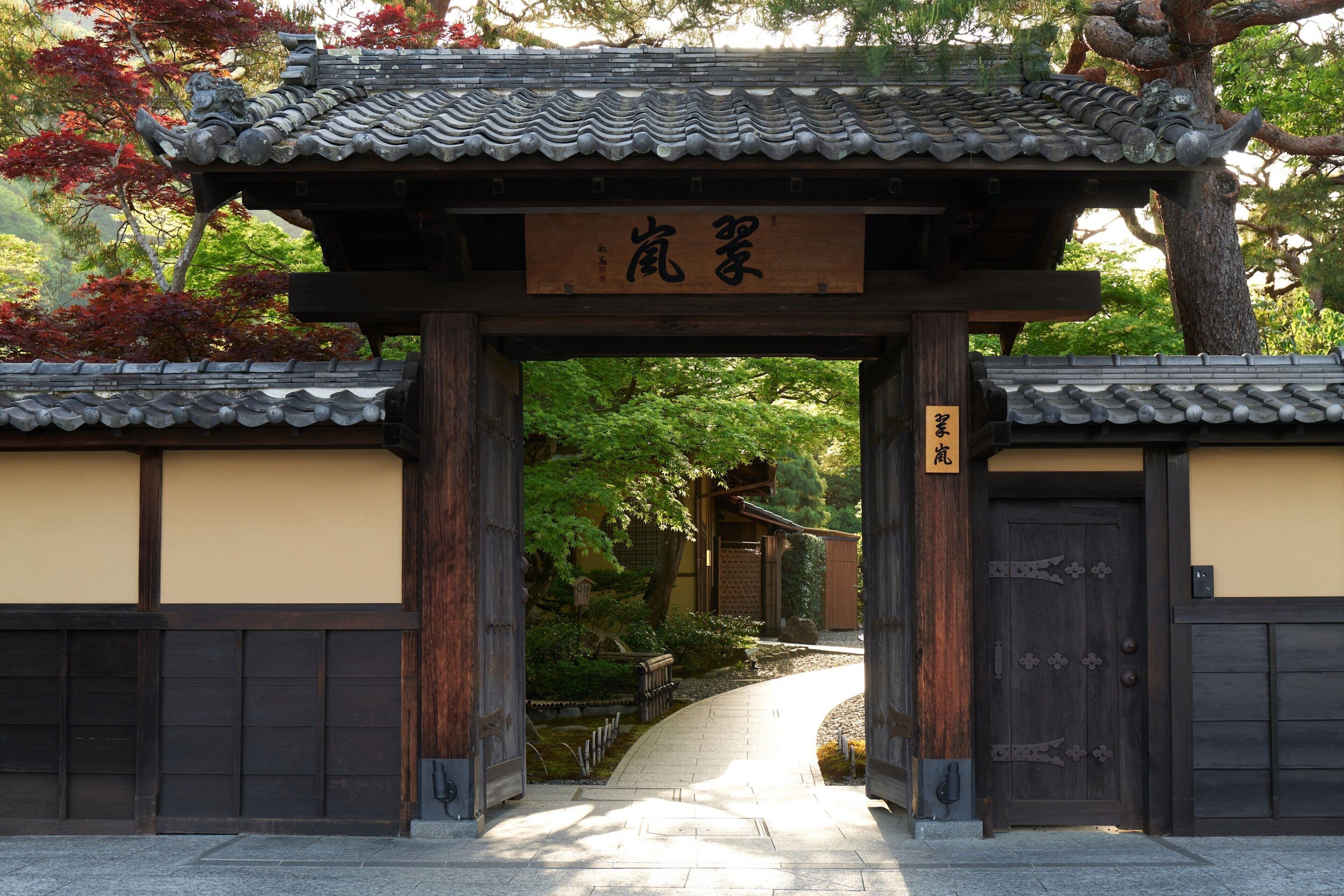 A traditional Japanese gate with a wooden structure and tiled roof, leading into a garden with a stone path.