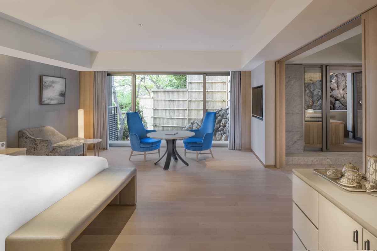 A modern hotel room with a large bed, a dining table with blue chairs, and a view of a bamboo fence and trees.